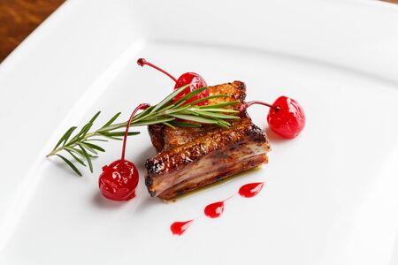 Grilled pork ribs decorated with rosemary and caramelized cherry served on a white plate in a restaurantの写真素材