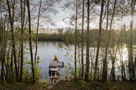 Aerial shot of woman in a yellow jacket relaxing on a wooden pier on a lake watching fall sunset alone. Enjoying nature, relaxation and meditationの写真素材