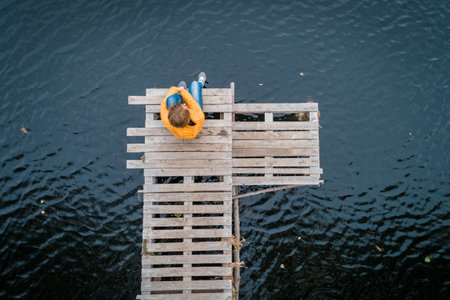 Aerial shot of woman in a yellow jacket relaxing on a wooden pier on a lake watching fall sunset alone. Enjoying nature, relaxation and meditationの写真素材
