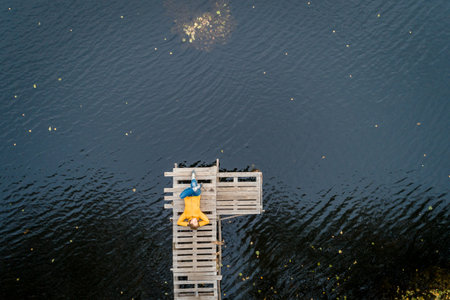 Aerial shot of woman in a yellow jacket relaxing on a wooden pier on a lake watching fall sunset alone. Enjoying nature, relaxation and meditationの写真素材