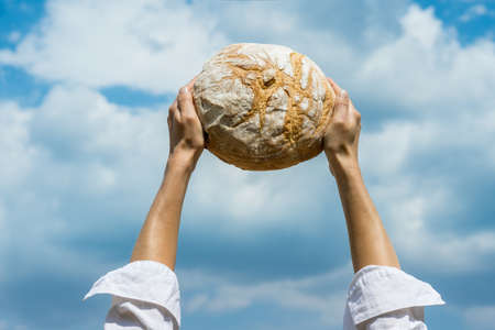Female hands holding home baked bread loaf above her head over a blue summer sky. World food security concept.の写真素材