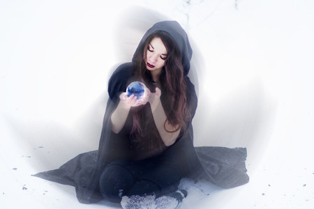 Witch or woman doing magic in black cloak with glass ball in white snow forestの写真素材