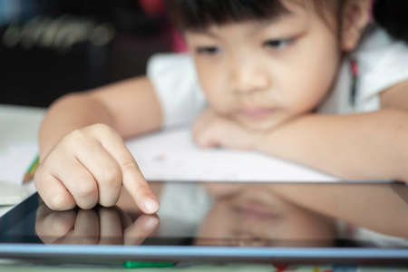 Closeup image of Child girl six years old touches the virtual screen of digital tablet.lonely, sad bored little girl sitting at the table in classroom.Selective focus on the child's finger.の写真素材