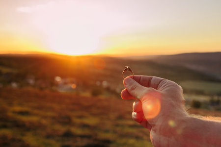 Man holding an engagement ring with a diamond in front of mountains and beautiful sunset. Proposal of marriage to a woman or a man in a beautiful place. Copy space for your text or advertisementの写真素材