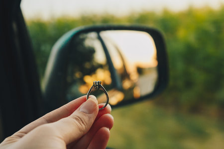 Woman holds engagement diamond ring in front of the side mirror with a view on the road in the meadow at sunset. Beautiful view from the car to the field. Travel concept, marriage proposal and adventures on the holidaysの写真素材