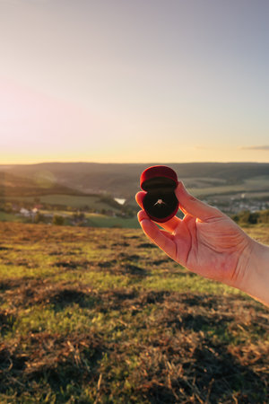Man holding a box with diamond engagement in front of mountains and beautiful sunset. Proposal of marriage to a woman or a man in a beautiful place. Copy space for your or advertisementの写真素材