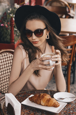 Beautiful girl in a hat sitting and sunglasses in a French cafe drinking coffee with a croissant. French cafe in city center, stylish outfit, atmospheric photoの写真素材