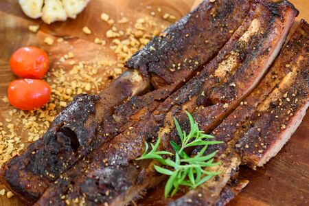 Baked pork ribs on a wooden plate decorated with garlic, tomatoes and green vegetablesの写真素材