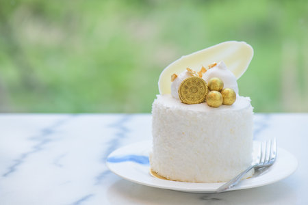 Coconut cake decorated with white chocolate, biscuits and gold leaf in a white plate on a granite table with a garden background.の写真素材