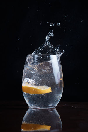 a slice of lemon falls into water in clear glass, splashes fly, citrus in glass with drink, dark background, close-upの写真素材