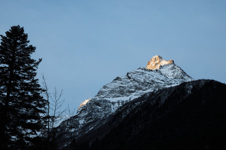 mountain peak at sunset, part of mountain is illuminated by rays of sun, clear sky and coniferous forest below, evening timeの写真素材