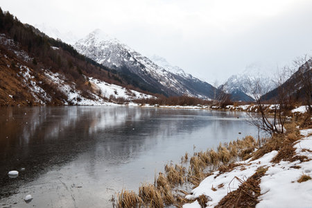 winter mountain lake in snow, in background of mountain, landscape pond with ice on waterの写真素材