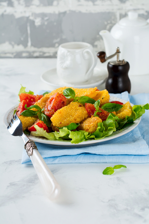 Fried cheese slices in corn grits, cooked tomatoes and lettuce on a light background. breakfast concept. Selective focus.の写真素材