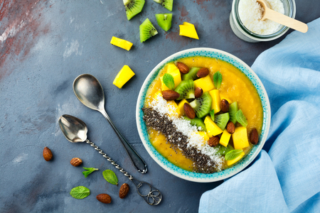 Mango smoothie bowl with pieces of kiwi, mango, coconut shavings and chia seeds on a stone background. Selective focus.の写真素材