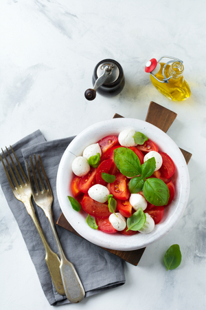 Traditional Italian caprese salad with tomatoes, mozzarella cheese and basil on a light marble background in a white old ceramic plate. Selective focus.Top view. Copy space.の写真素材