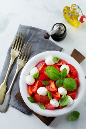 Traditional Italian caprese salad with tomatoes, mozzarella cheese and basil on a light marble background in a white old ceramic plate. Selective focus.Top view. Copy space.の写真素材