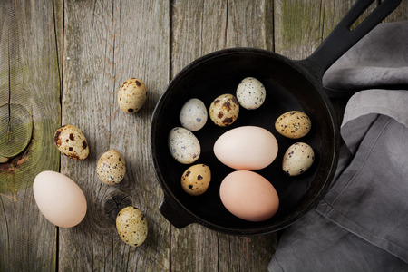 Whole quail and chicken eggs in a small cast-iron frying pan on the background of an old wooden texture. Selective focus. Top view with spaceの写真素材