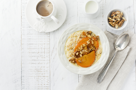 Oatmeal porridge with caramelized persimmon and walnuts in a light ceramic bowl on a white wooden background. Selective focus. Top view. Copy space.の写真素材