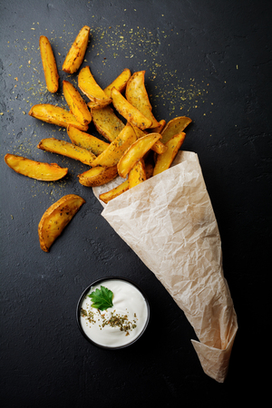 Baked potatoes with greens and sauce in paper bag, scattered on dark concrete or ceramic background. Selective focus. View from above. Place for text.の写真素材