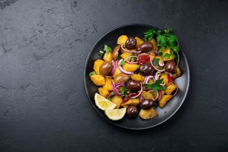 Warm potato salad with olives, pepper, parsley and red onion on black ceramic plate on dark concrete background. Selective focus. Top view. Copy space.の写真素材