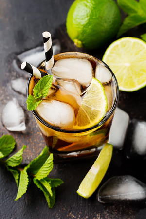 Summer cooling alcoholic beverage from cola, whiskey, lime and liqueur in glass on dark concrete old background.の写真素材