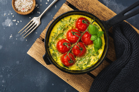 Frittata with arugula, potatoes and cherry tomatoes in iron pan on old stone table background. Top...