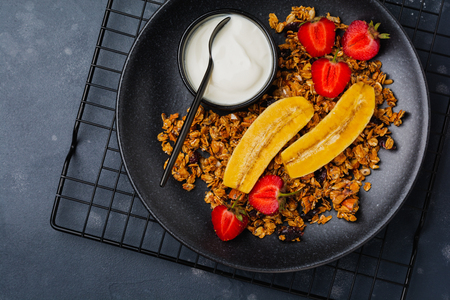 Oatmeal granola with yoghurt, fresh strawberries and banana, chia seeds, sunflower and honey in black ceramic plate on dark background. Top view.の写真素材