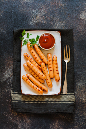 Grilled BBQ sausages with sauce and ketchup and garlic on ceramic plate on dark concrete background. Top view.の写真素材