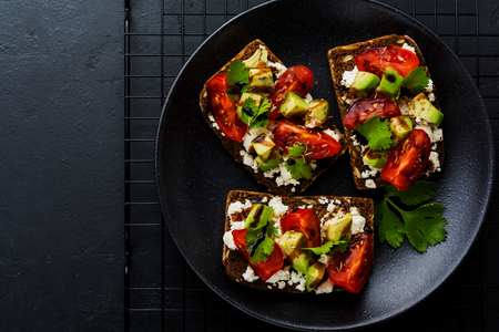 Avocados, cherry tomatoes and feta cheese sandwiches with balsamic sauce and cilantro on old olive board on dark black background. Top view.の写真素材