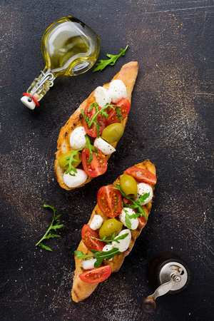 Caprese bruschetta toasts with cherry tomatoes, mozzarella, olives and basil on old dark background. Top view.の写真素材