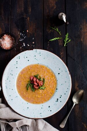 Homemade red lentil cream soup with bacon and arugula in a white plate on an old wooden background. Rustic style. Top view.の写真素材