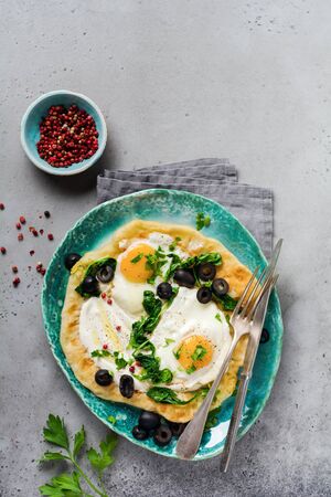 Turkish eggs flatbread with yogurt, cheese, olives, spinach and red pepper on ceramic vintage plate on gray old background. Top view.の写真素材