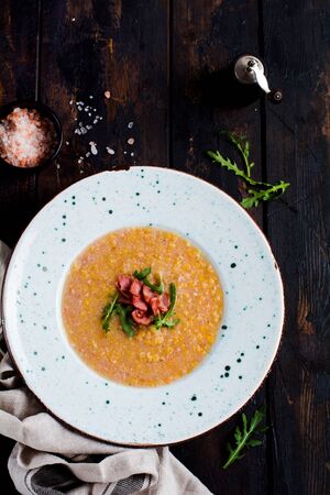 Homemade red lentil cream soup with bacon and arugula in a white plate on an old wooden background. Rustic style. Top view.の写真素材