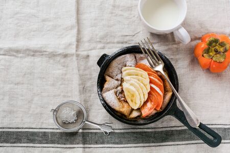 "Dutch baby" pancake with apple, persimmon, banana, cinnamon in small iron pan on wooden table background. Top view.の写真素材