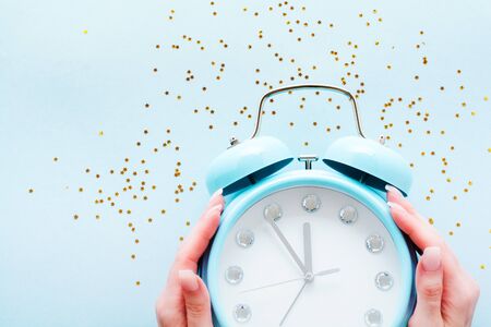 Female hands holding big blue alarm clock lying and surface showing 23 hours 55 minutes with shiny...