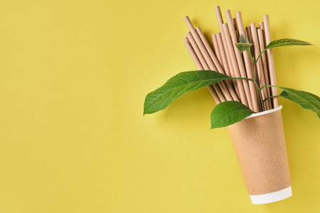 Drinking tubes made of paper and cornstarch, biodegradable material and eco paper glasses with green sprout leaves on yellow trend color 2021 background. Zero waste and plastic free concept. Top view.の写真素材