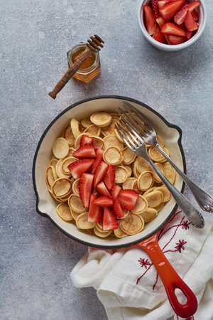 Mini white pancake cereal with strawberries in frying pan for breakfast on gray background. Trendy home breakfast with tiny pancakes. Top view.の写真素材