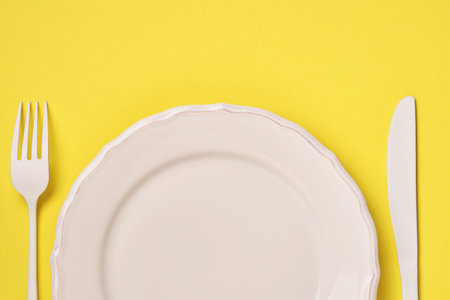 White plate, fork and spoon on a trendy yellow background. Time to eat. Diet concept. Top view.の写真素材