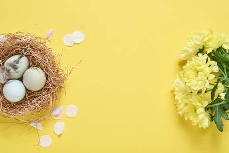 Easter light blue eggs in white vintage metal holder with feathers, ribbon, yellow chrysanthemums flowers and blank paper for text on yellow background. Mock up. Spring Happy Easter holiday card.の写真素材