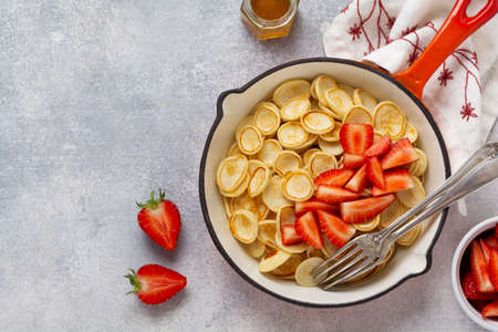 Mini white pancake cereal with strawberries in frying pan for breakfast on gray background. Trendy home breakfast with tiny pancakes. Top viewの写真素材