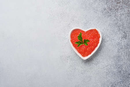 Red caviar in bowl in shape of heart served with lemon and ice cubes on gray background table. Space for text.の写真素材