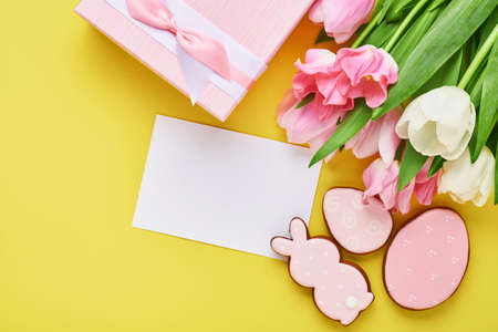 Easter greeting card with easter gingerbread eggs gift box and tulip flowers over yellow background....