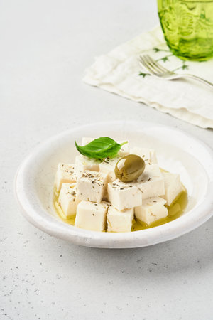 Feta cheese cubes with rosemary, olives and olive oil sauce in white bowl on light gray background. Traditional Greek homemade cheese. Selective focus.の写真素材