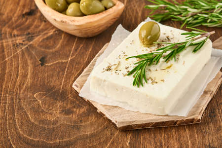 Cheese feta with rosemary, herbs, olives and olive oil on wooden cutting board on old wooden background. Traditional Greek homemade cheese. Selective focus.の写真素材