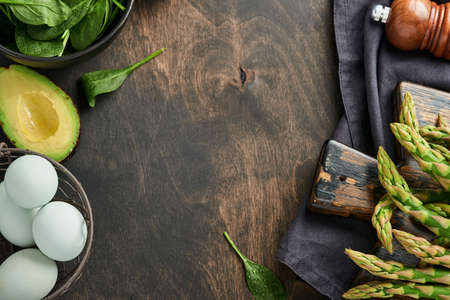 Bunch of fresh green organic asparagus spinach, avocado, chicken eggs and pepper seasoning on black old wooden background, top view. Food cooking background with copy space. Mock up.の写真素材
