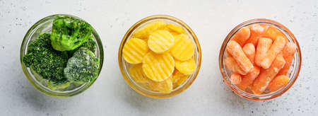 Frozen vegetables such as green broccoli, yellow and baby carrot in glass bowls on ice and concrete gray background with copy space. Banner.の写真素材