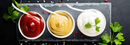 Set of three sauces - mayonnaise, mustard and ketchup in white ceramic bowls on black stone or...