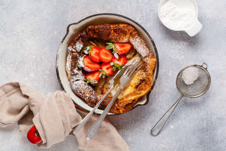 Dutch baby pancake with fresh strawberry berry and sprinkled with icing sugar powder in red pan on white kitchen background. Top viewの写真素材