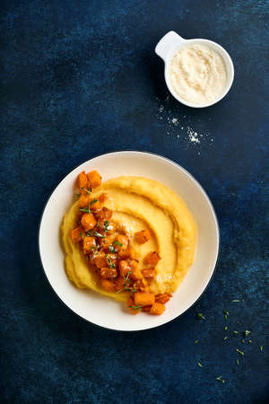 Polenta with butter, pumpkin, garlic, savory and parmesan cheese in white bowl on dark blue concrete background. Traditional Basic Italian food, vegan food. Top view. Copy space.の写真素材