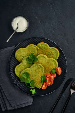 Spinach avocado pancakes in the shape of a heart with greek yogurt sauce and cherry tomatoes on a black plate over dark background. Breakfast food concept. Top view with copy space.の写真素材